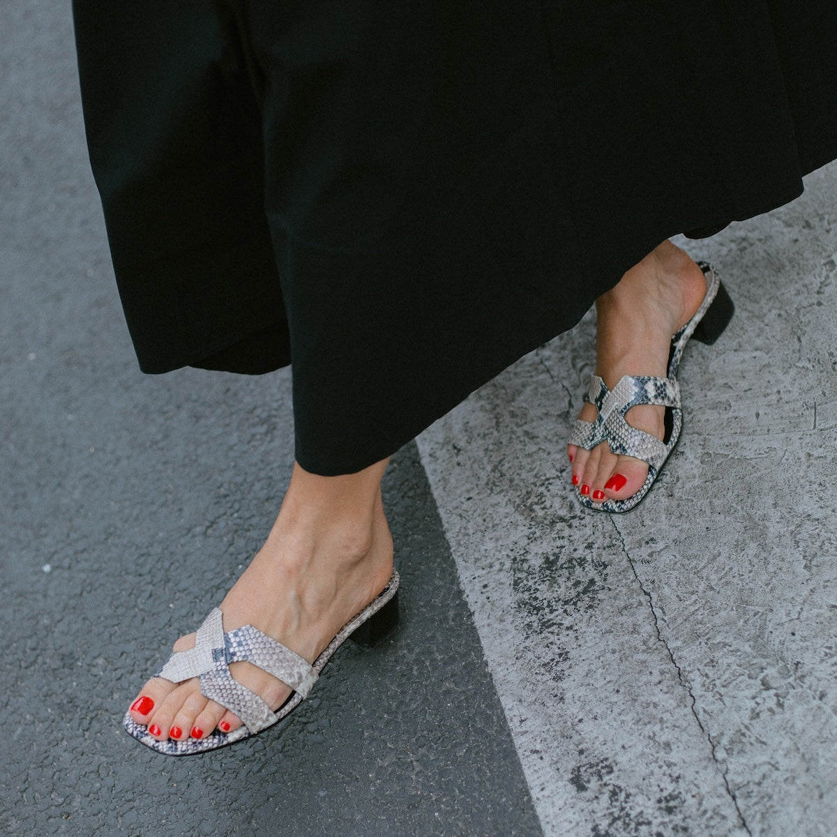 A person wearing a long black skirt and open-toed margaux-development The MX 35 - Natural Python Embossed sandals with block heels and red nail polish stands elevated on the edge of a street.