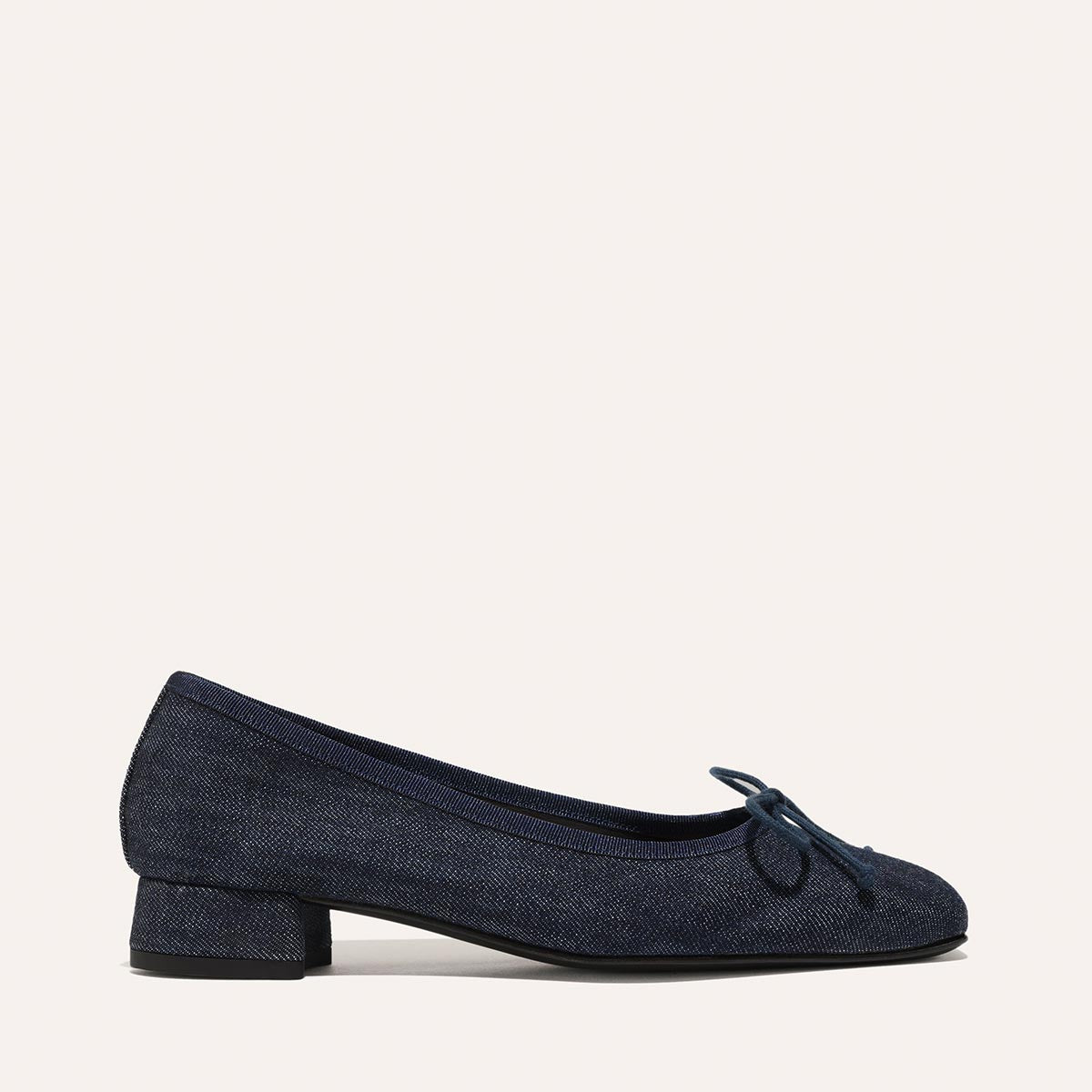 The Ada - Indigo Denim by margaux-development is a classic dark blue denim ballet pump with a low block heel and small bow on the rounded toe, displayed on a light background.