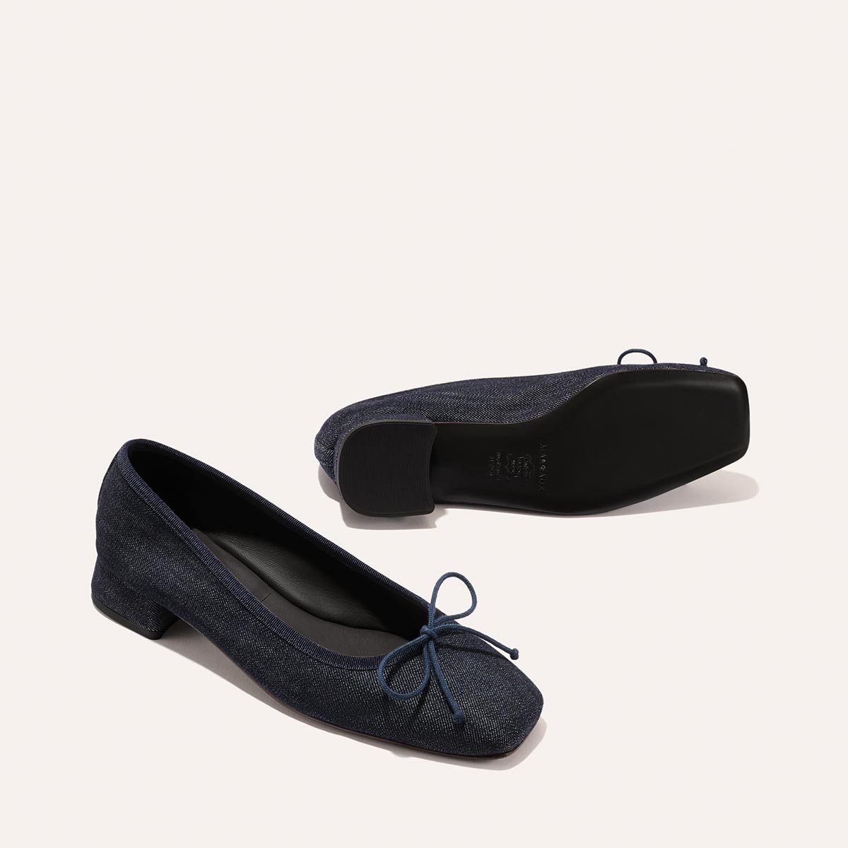 The Ada – Indigo Denim ballet pumps by margaux-development feature dark blue denim, low block heels, and small front bows. One shoe stands upright while the other lies on its side, both set against a plain light background.