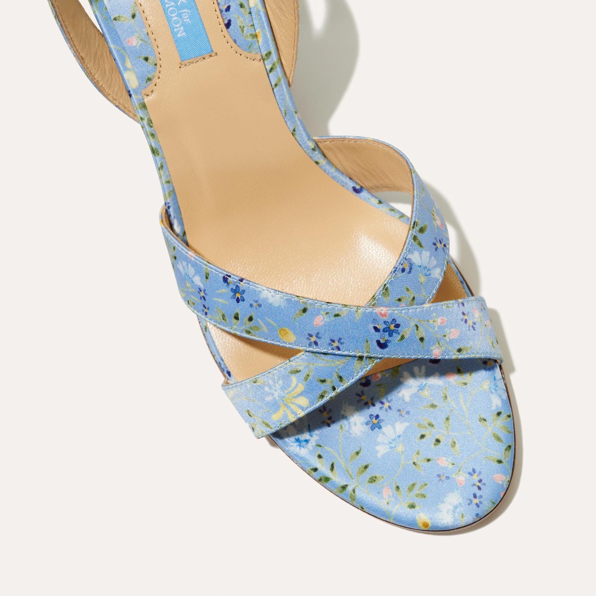 A close-up of margaux-developments The City Sandal - Blue Floral Satin shows heeled sandals with crossed straps in light blue floral satin and a cushioned insole for all-day comfort, set against a plain background.