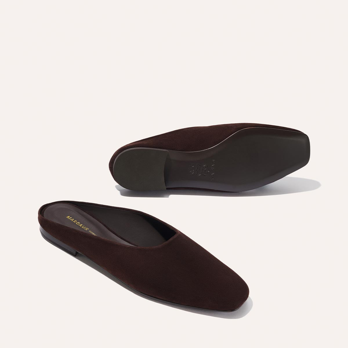 The Clara - Espresso Suede by Margaux: brown suede slip-on shoes with flat soles and a slipper-like shape, shown on a light background. One shoe stands upright, the other displays its sole.