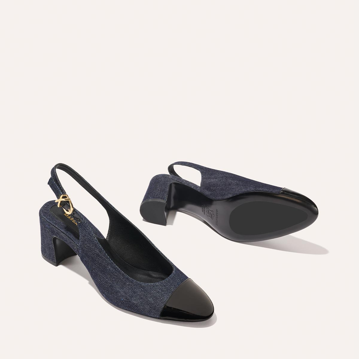 The Cluny Slingback by margaux-development features indigo denim and patent, block heels, black pointed toe caps, and gold buckle details. Displayed on a light background, one shoe stands upright while the other lies on its side.