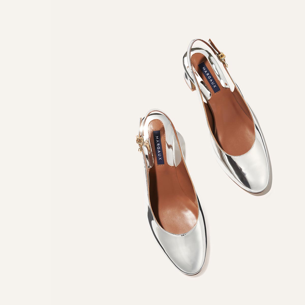 The Cluny Slingback - Silver Mirror by Margaux, with its brown insoles and gold buckle detail, exudes Parisian elegance and New York sensibility in a chic, shiny silver finish—perfectly photographed on a light background.