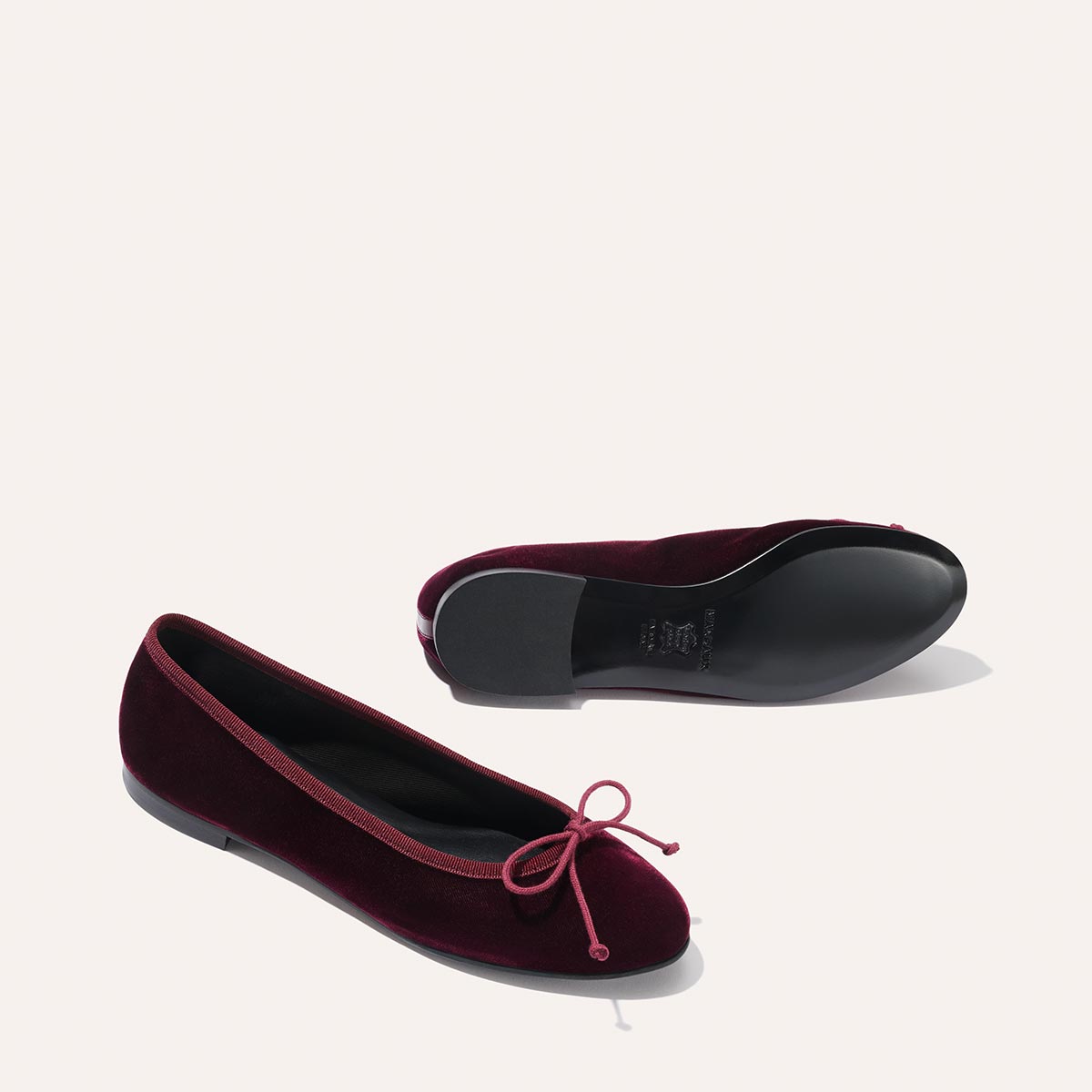 A pair of The Demi - Burgundy Velvet ballet flats by margaux-development, featuring matching bows; one shoe displays the top view upright, while the other lies on its side revealing the black sole.