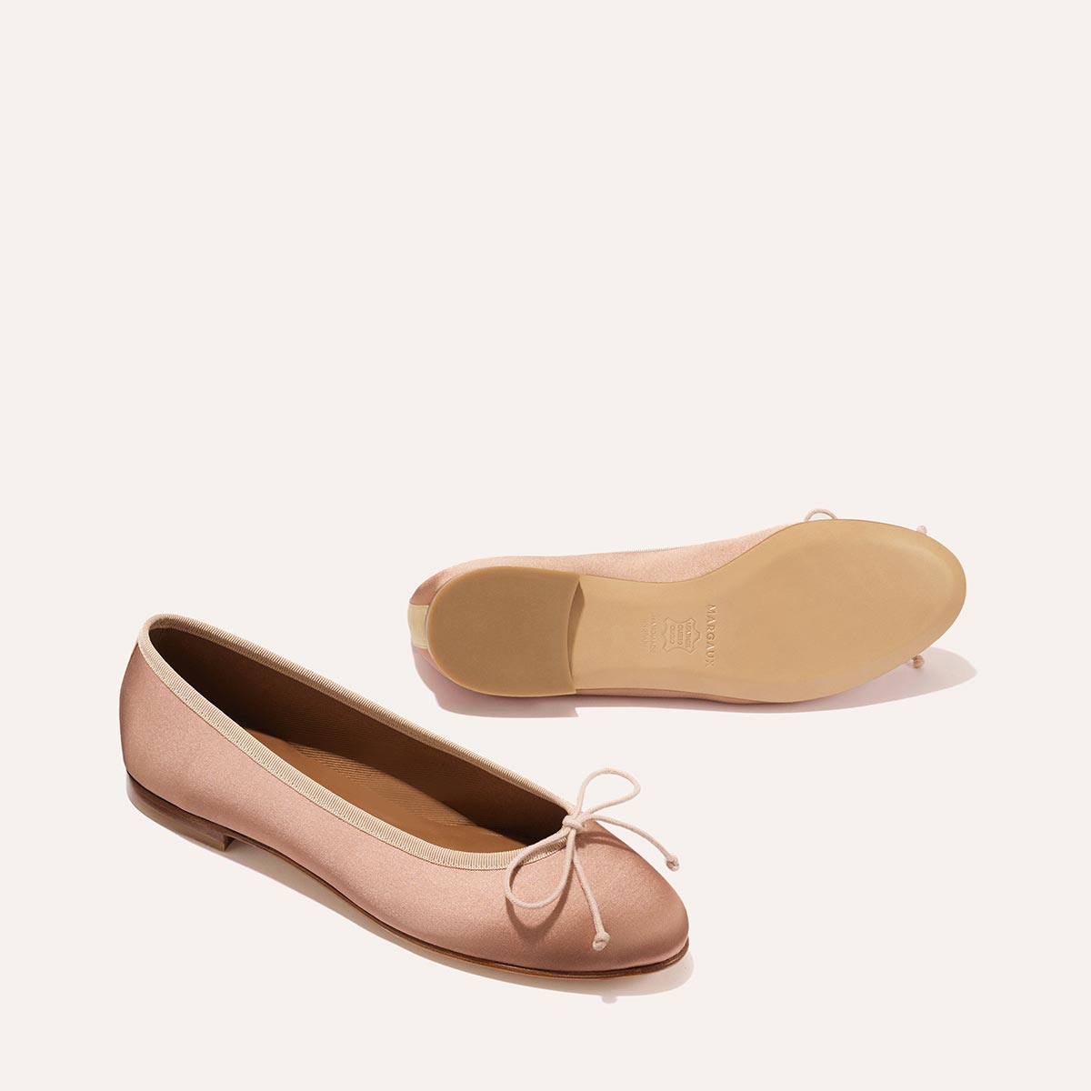 The Demi - Dusty Pink Satin by margaux-development features light pink ballet flats with a small toe bow; one stands upright, the other rests sideways showing the sole. Handmade in Spain for timeless, comfortable style.