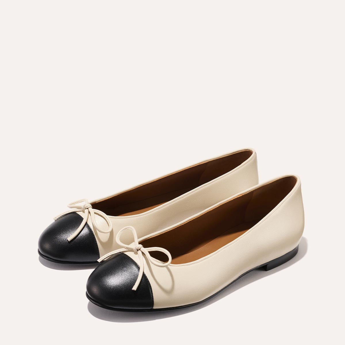 The Demi - Ecru and Black Nappa by margaux-development: womens ballet flats, handmade in Spain, with cream bodies, black rounded toe caps, and cream bows, displayed on a plain light background.
