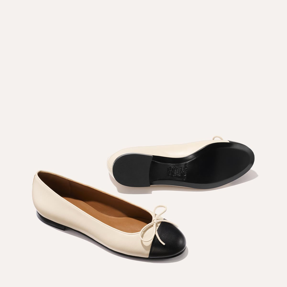 The Demi - Ecru and Black Nappa by margaux-development are bestselling handmade Spanish ballet flats in ivory with black capped toes, bow accents, tan insoles, and black soles—shown on a light background in two positions.