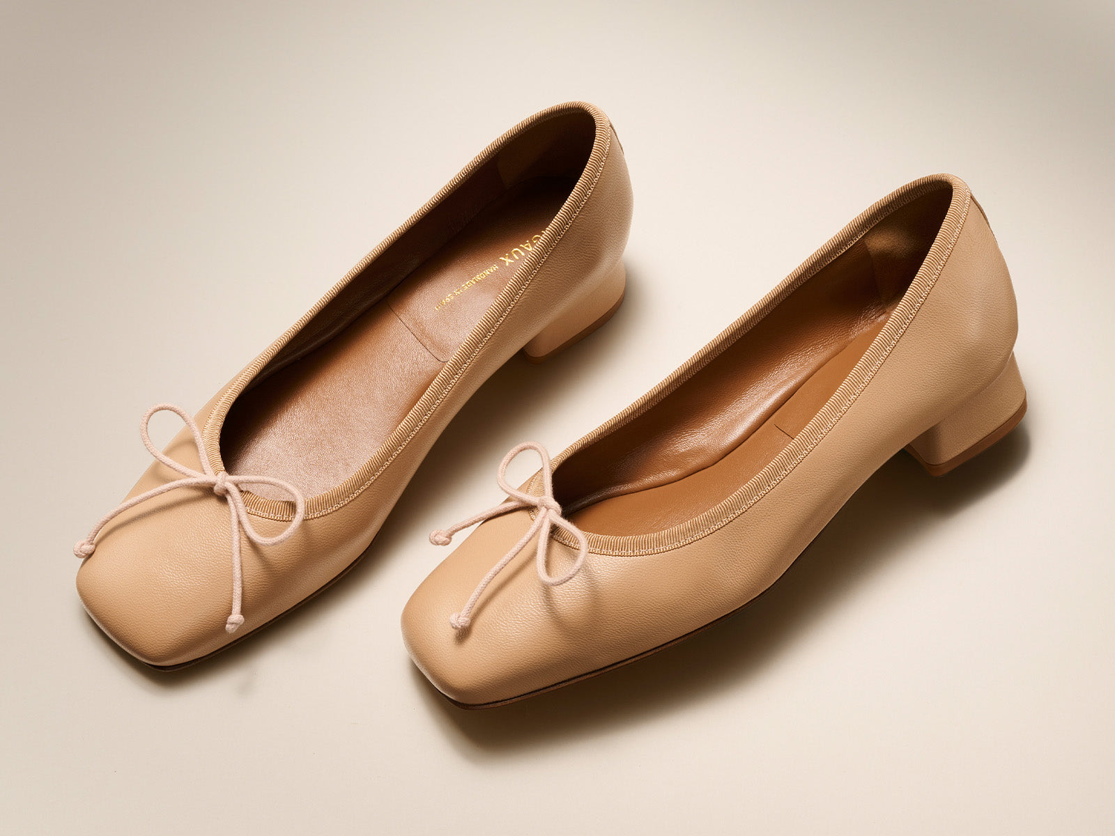 A pair of beige leather ballet pumps with square toes, low block heels, and small bows on the front, displayed on a light, neutral background.