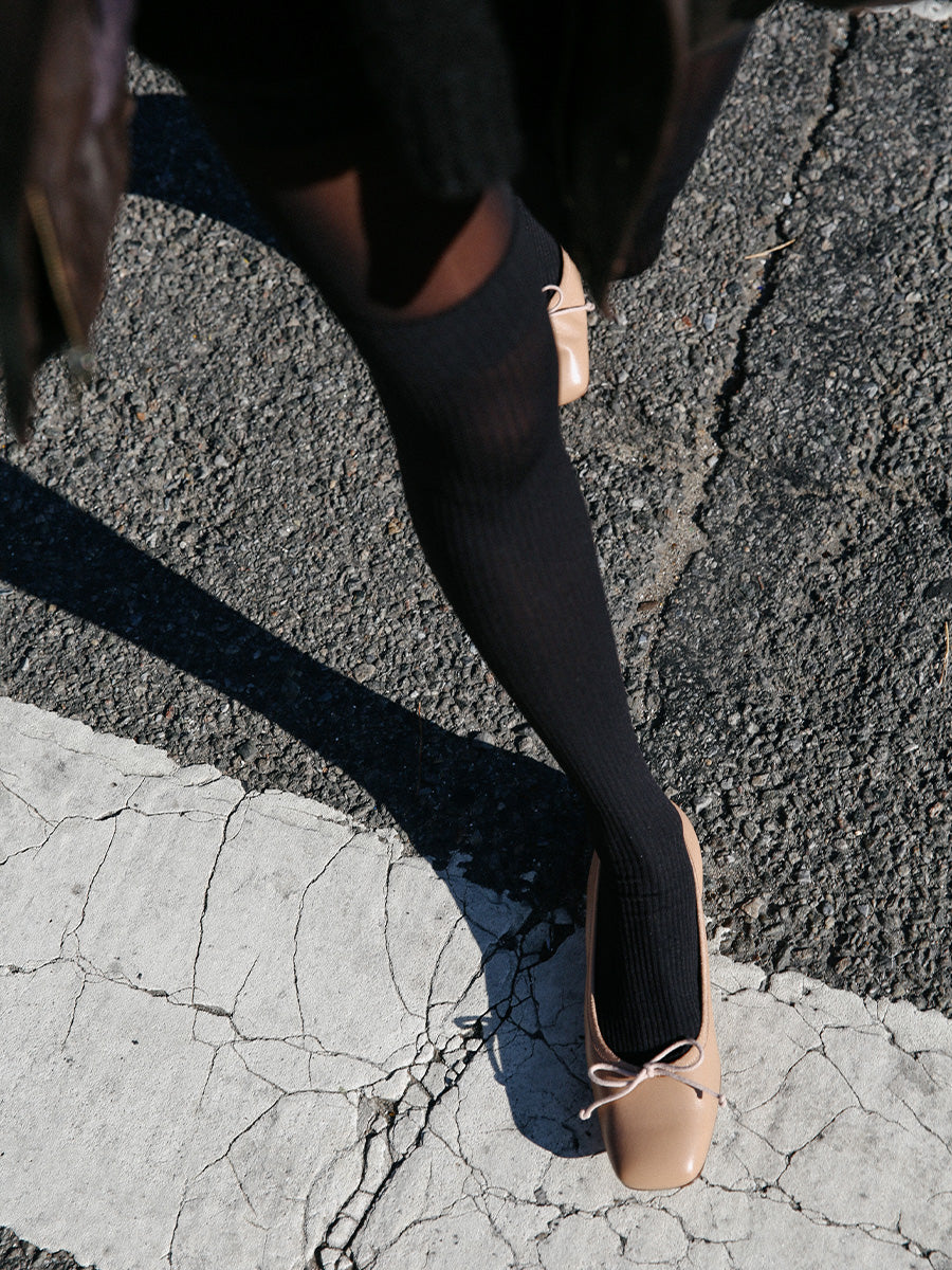 A person wearing black knee-high socks and beige ballet flats steps on a cracked crosswalk line on an asphalt road.