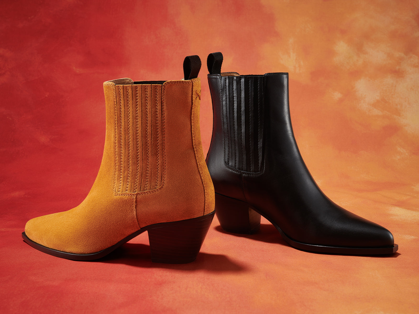 Two ankle boots are displayed side by side against an orange background: one boot is tan suede, and the other is black leather, both with chunky heels and elastic side panels.