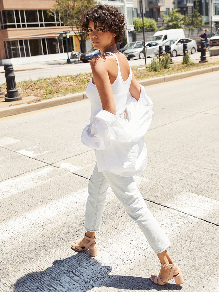A woman with curly hair, wearing a white tank top, white pants, and nude heels, walks across a city street while looking over her shoulder. She has a white shirt draped over her arms.