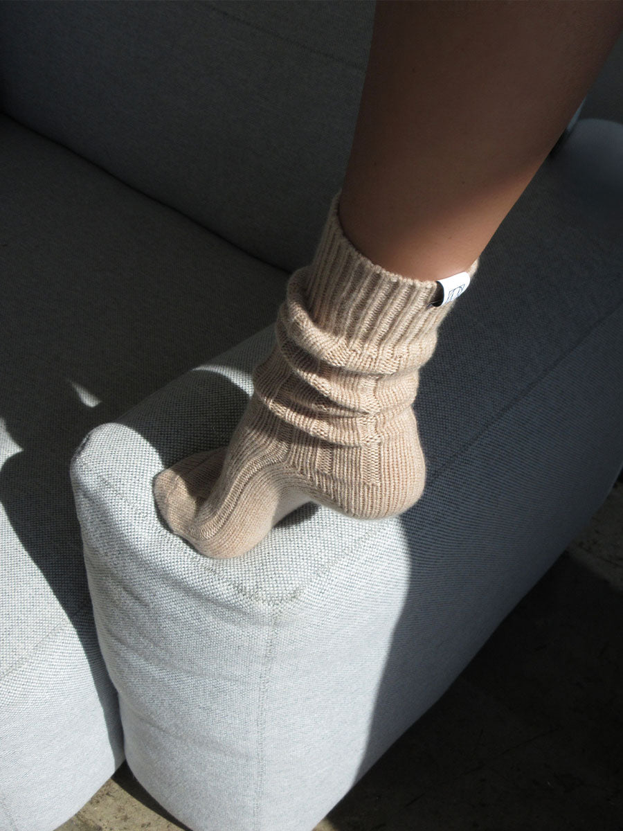 A person wearing beige, ribbed socks rests their foot on the armrest of a light gray upholstered sofa. Sunlight casts shadows on the furniture and foot.