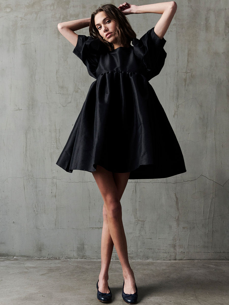 A woman stands against a bare concrete wall, wearing a short, loose-fitting black dress with ruffled sleeves and black heels. She poses with her hands touching her head and her legs crossed at the ankles.