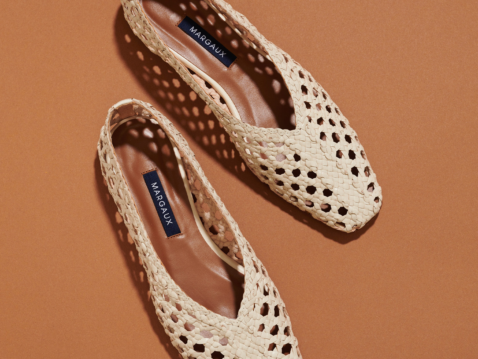 A pair of woven, beige slingback flats with pointed toes, featuring a cutout design and dark insoles labeled Margaux, placed on a brown background.