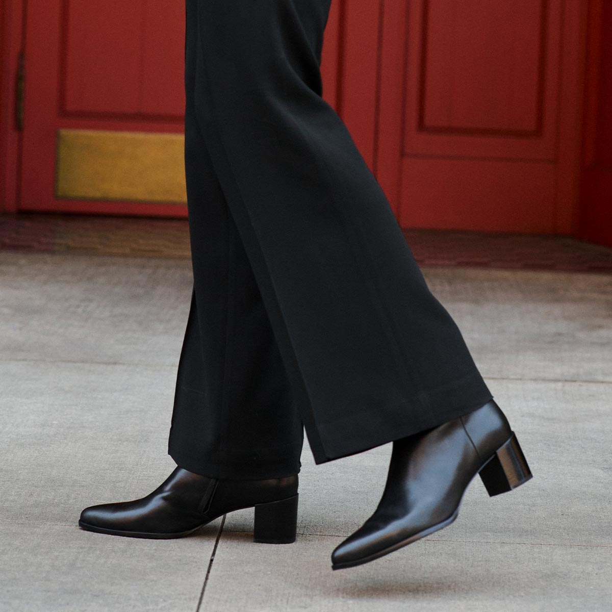 A person wearing Margaux's The Downtown Boot - Black Calf with a pointed toe and black pants walks on a sidewalk, with a red door in the background.