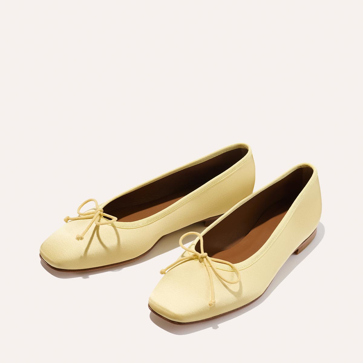 The Fonteyn - Butter Satin by Margaux: handcrafted light yellow ballet flats with square toes, small front bows, and brown insoles, designed for comfort and displayed on a plain white background.