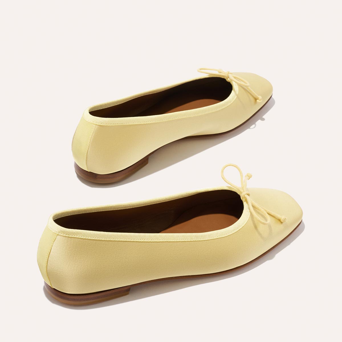 The Fonteyn - Butter Satin by Margaux: handcrafted light yellow ballet flats with rounded toes, small front bows, and brown flat soles, displayed on a plain light background.