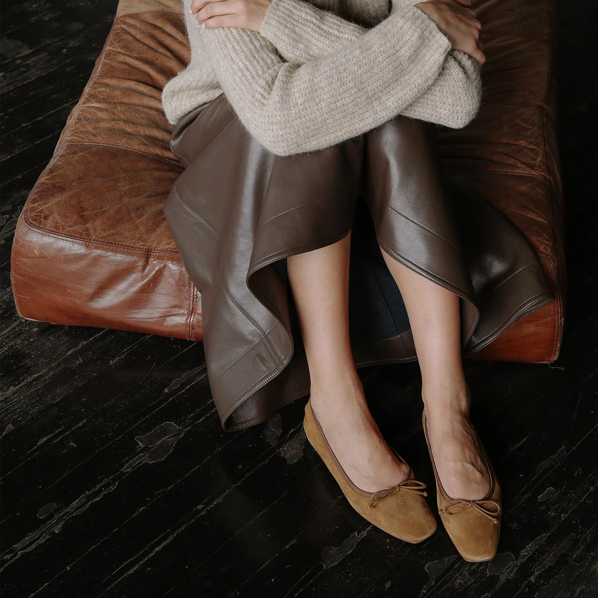 A person sits on a brown leather seat with arms crossed, wearing a beige sweater, brown leather skirt, and Margaux’s The Fonteyn - Cedar Suede ballet flats, handcrafted and tan with bows, on a dark wooden floor.