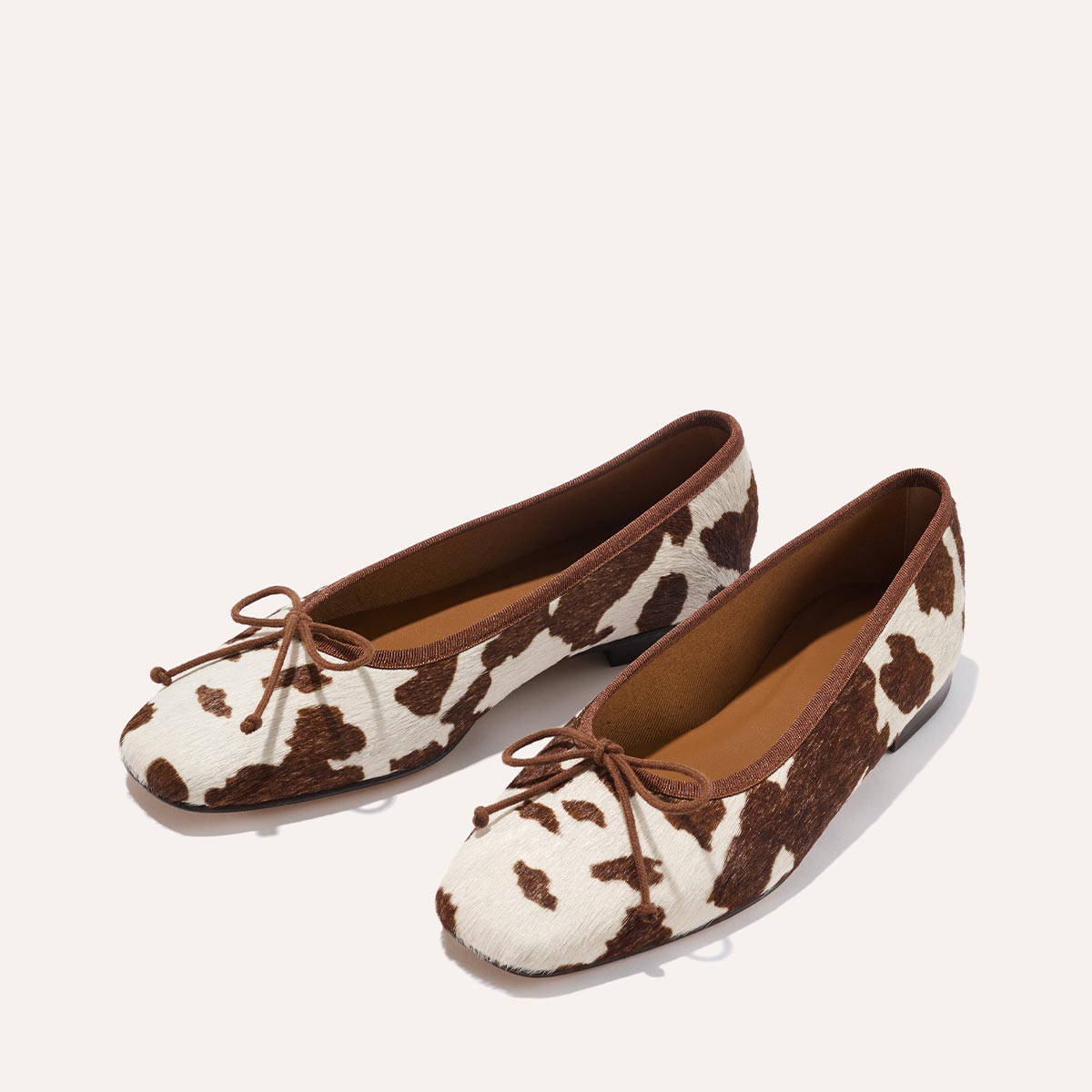 The Fonteyn - Cow Print Haircalf by Margaux: stylish ballet flats with brown cow-print pattern, brown trim, and small front bows, shown on a light background.