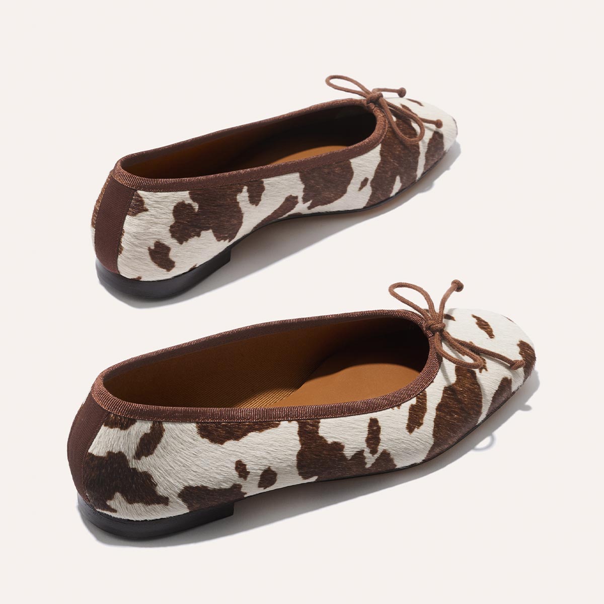 The Fonteyn - Cow Print Haircalf by Margaux: stylish ballet flats in brown and white cow print with brown trim, front bow detail, and black soles, shown on a light background.