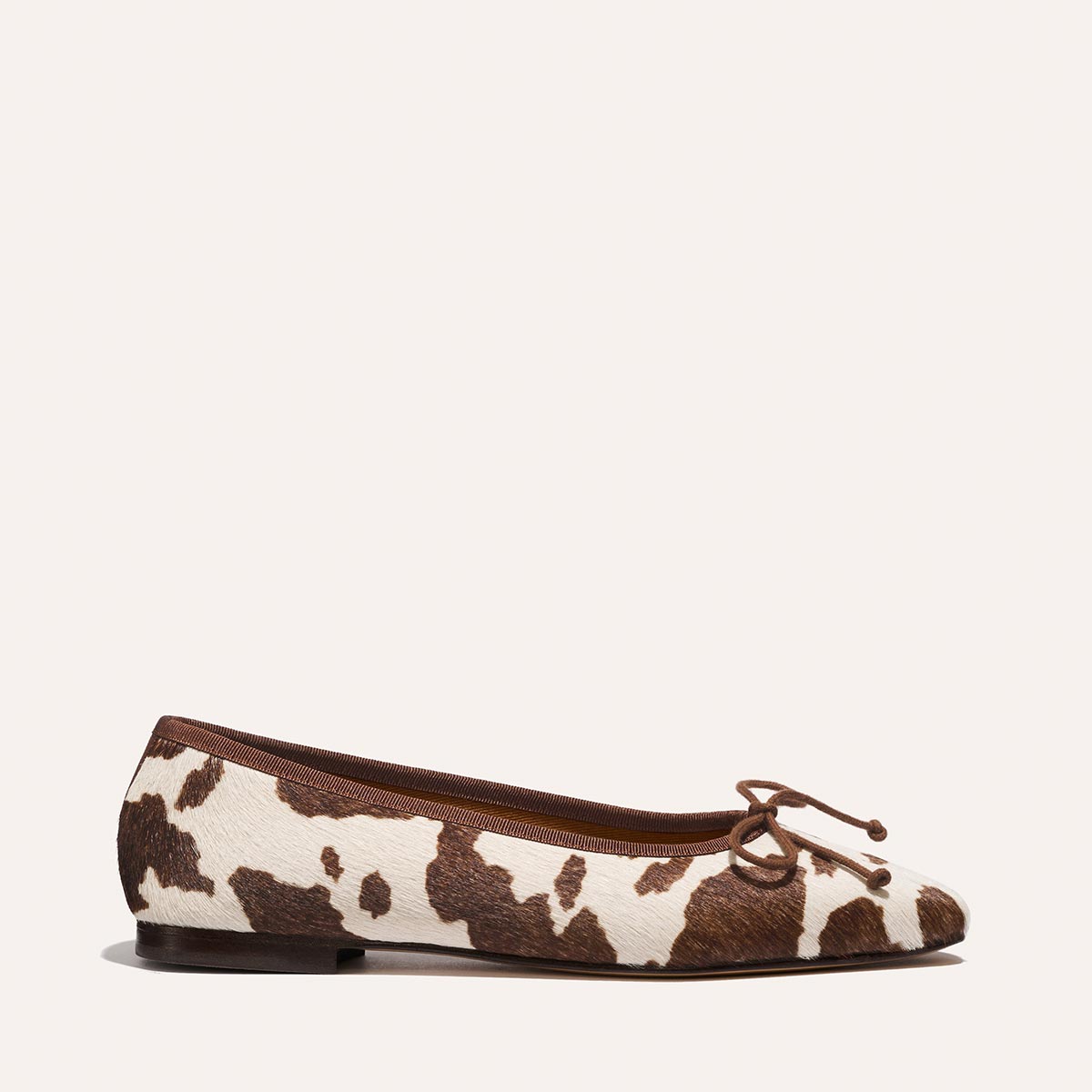 The Fonteyn - Cow Print Haircalf by Margaux is a chic ballet flat with a brown and white cowhide pattern, brown trim, a small toe bow, and a slim black sole, shown side-on against a plain light background.