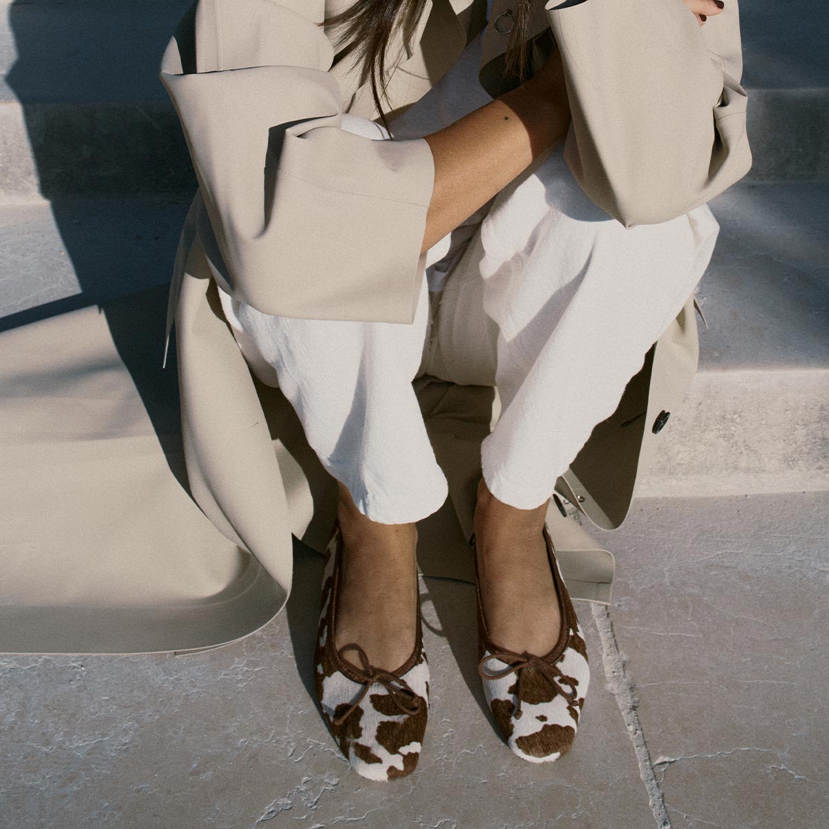 A person sits on concrete steps wearing a beige coat, white pants, and Margaux's The Fonteyn - Cow Print Haircalf ballet flats. Their arms are crossed as sunlight casts shadows over the comfort-first scene.