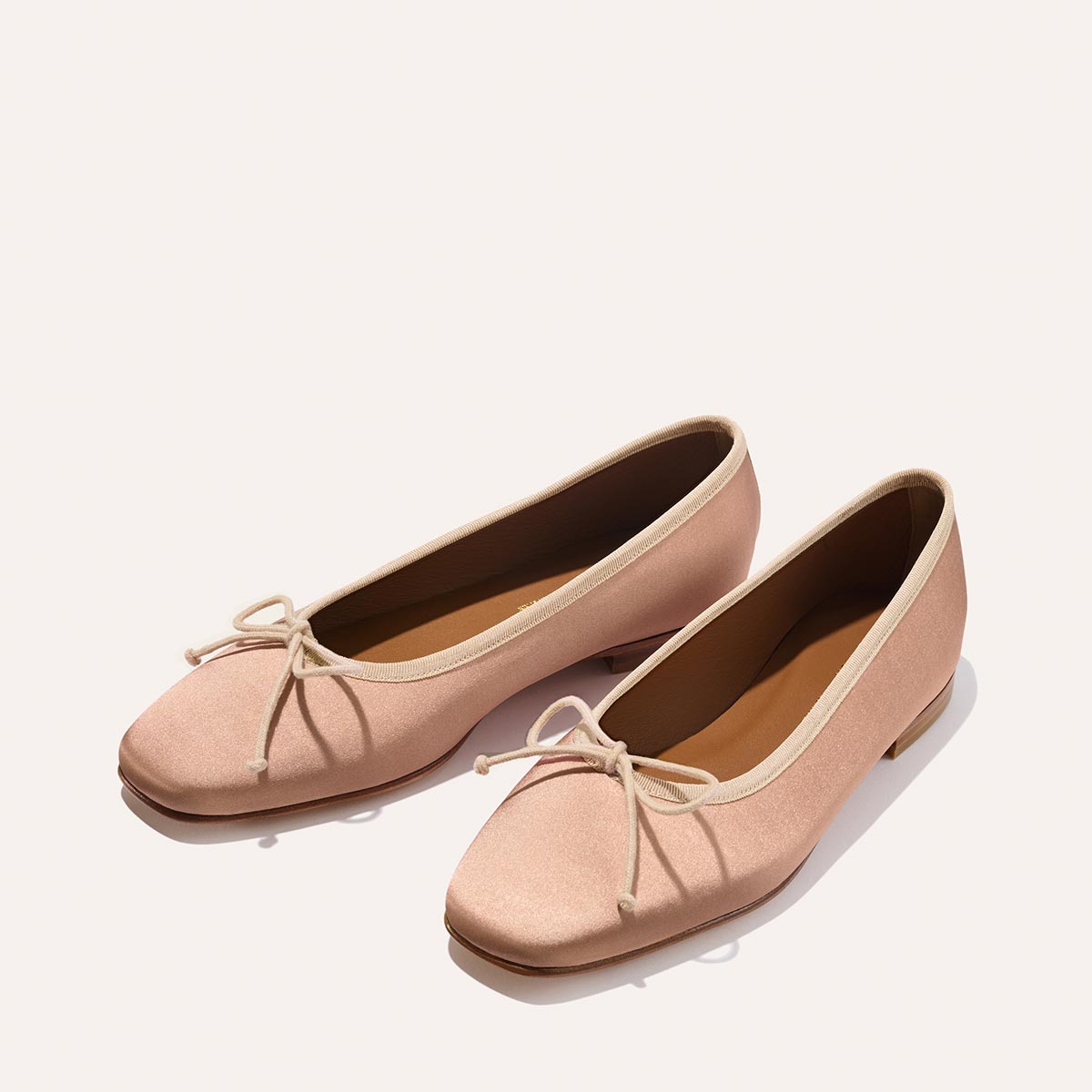 The Fonteyn - Dusty Pink Satin by Margaux: these light pink ballet flats feature small front bows, tan insoles, and flat soles, designed for comfort and shown against a plain light background.
