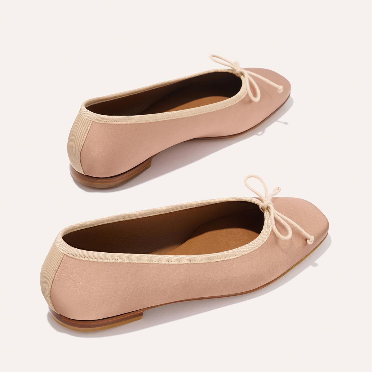 The Fonteyn - Dusty Pink Satin by Margaux: pink ballet flats with tan trim, small front bows, and tan soles, handcrafted for comfort-first fit. Displayed on a light background.