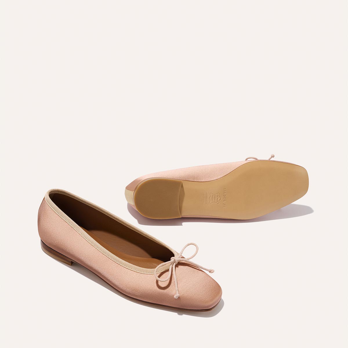 The Fonteyn - Dusty Pink Satin by Margaux: Handcrafted ballet flats in light pink satin, featuring small toe bows. One shoe stands upright while the other displays the sole on a plain, light background for a comfort-focused look.