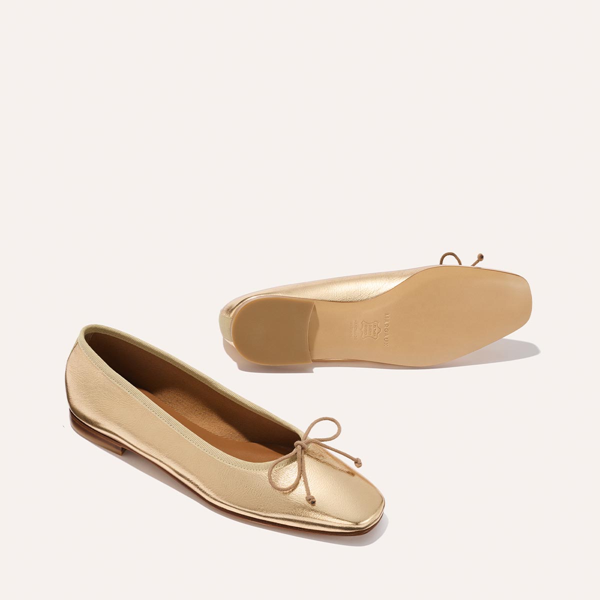 The Fonteyn - Gold Crinkle Nappa by Margaux: a pair of gold ballet flats with a bow accent. One shoe stands upright while the other shows off its smooth sole, set against a light, minimalist background.