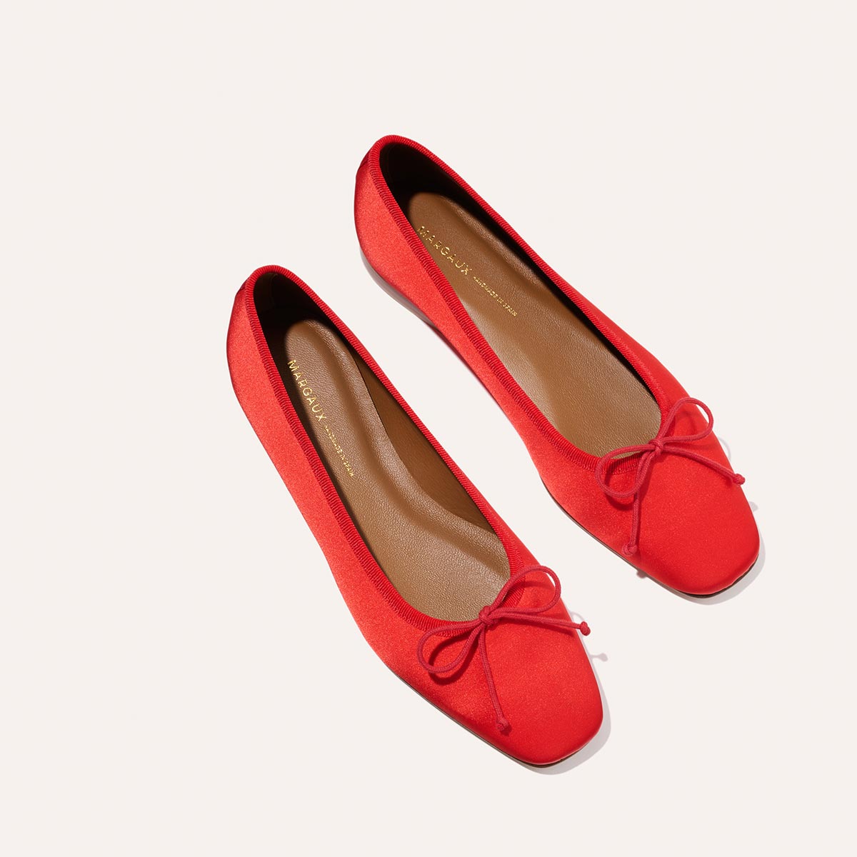 The Fonteyn - Poppy Satin by Margaux: bright red handcrafted ballet flats with square toes, red bows on top, and tan insoles, displayed on a light background.