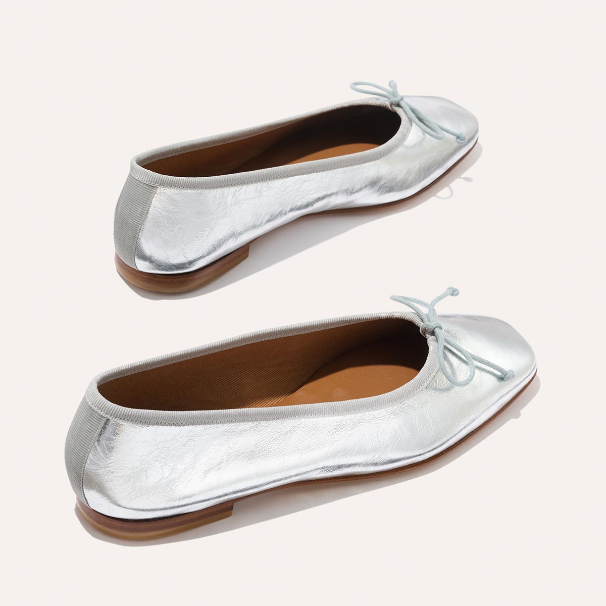 The Fonteyn - Silver Crinkle Nappa by Margaux: silver ballet flats with light blue bows, brown soles, and a beige inner lining, displayed on a plain light background.