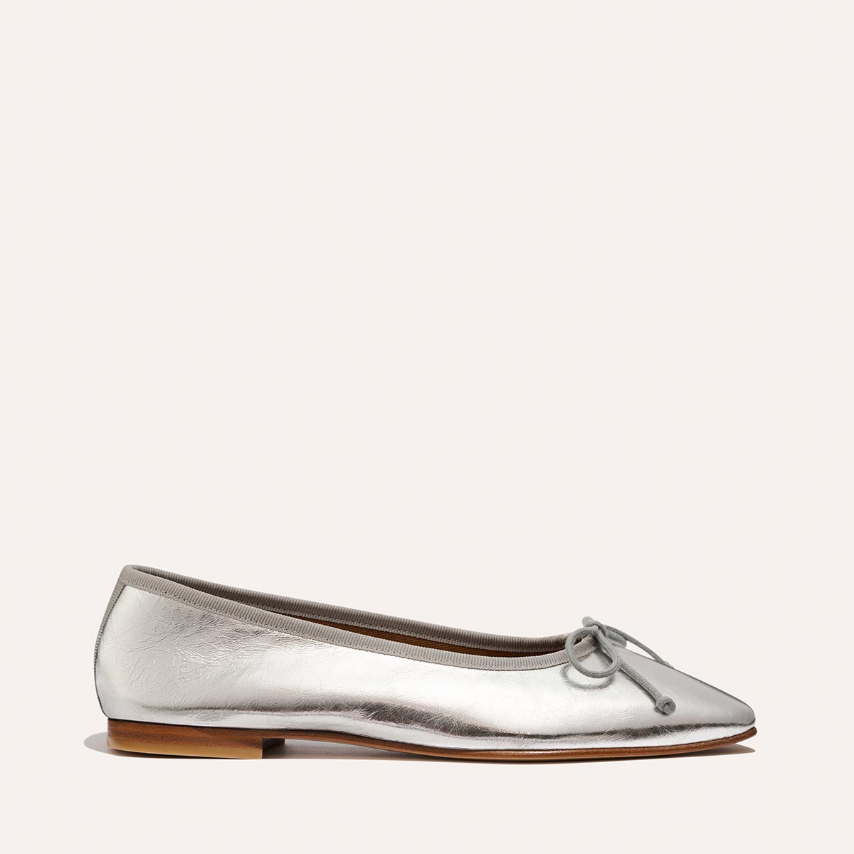 The Fonteyn - Silver Crinkle Nappa by Margaux is a silver ballet flat featuring a small bow on the rounded toe, beige trim, and a low wooden heel, shown against a plain light background.