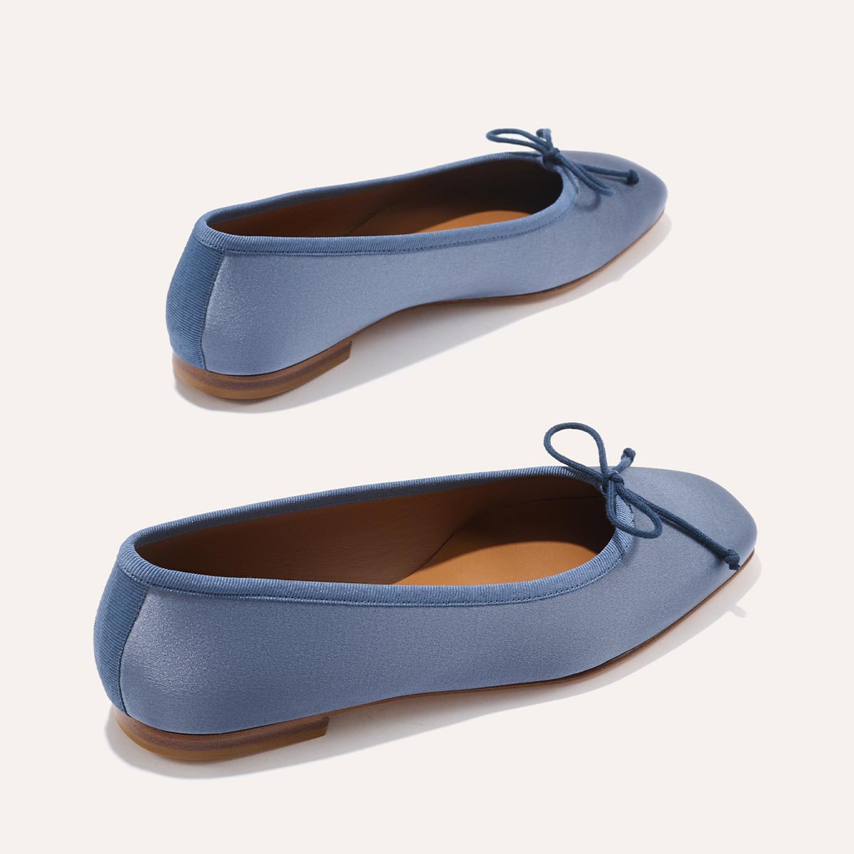 Two Margaux The Fonteyn ballet flats in slate blue satin, featuring small bows and tan soles, are displayed on a light background to showcase their side and back views.