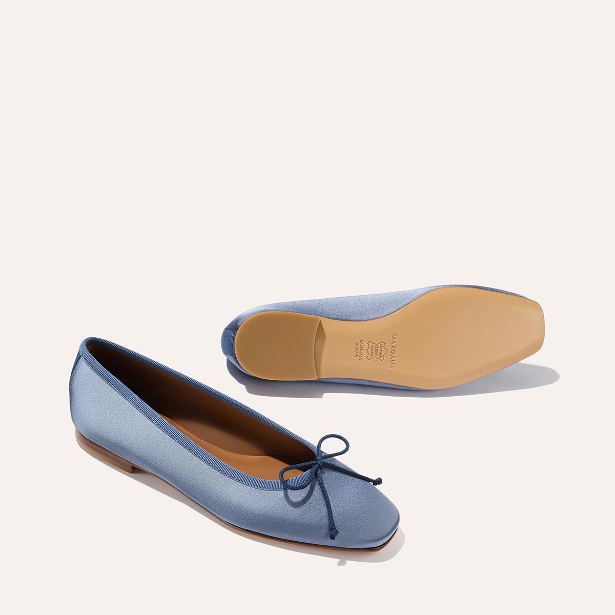 The Fonteyn - Slate Blue Satin ballet flats by Margaux, shown with one shoe in side view and the other from the bottom, feature light blue satin and small front bows against a clean, light background.
