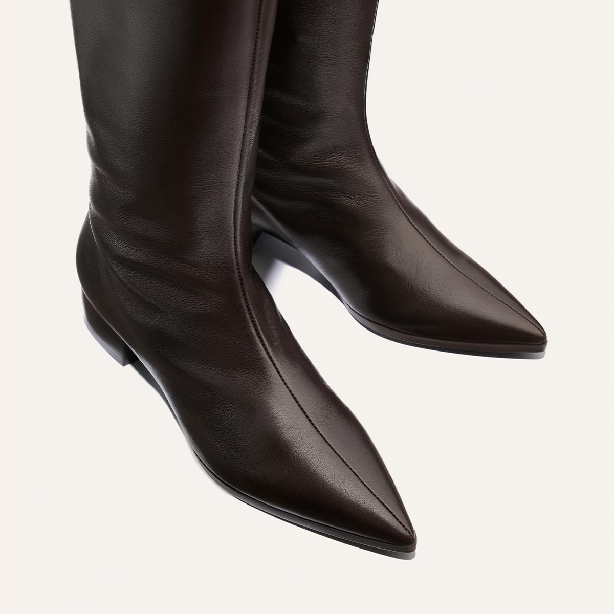 A close-up of The Hayes Knee-High Boot in Dark Chocolate Calf by Margaux, handcrafted from supple leather with pointed toes and low heels, displayed on a light background. These comfortable, handmade boots offer timeless style and enduring comfort.
