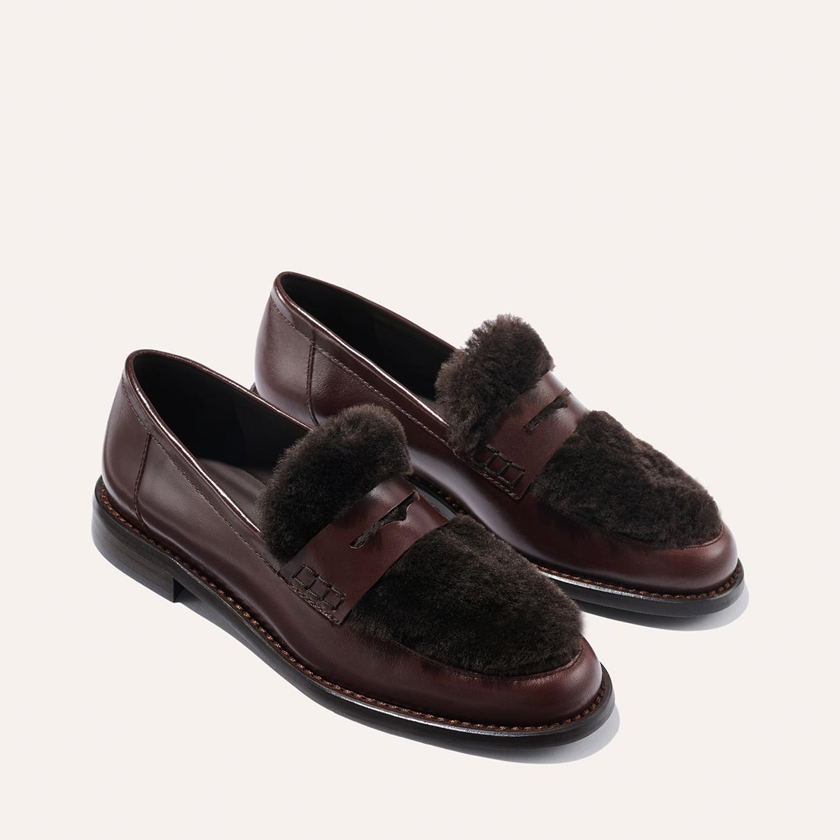 The Marlowe Loafer by Margaux in Mahogany Calf and Shearling features stitched accents, dark brown fur detailing, and black rubber soles, shown on a light background.