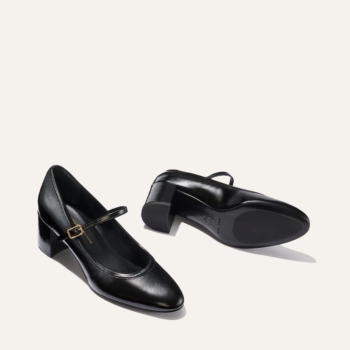 The Mira Jane - Black Crinkle Patent by margaux-development features round toes, low block heels, and thin gold-buckled straps. One shoe stands upright while the other lies on its side, revealing the sole.