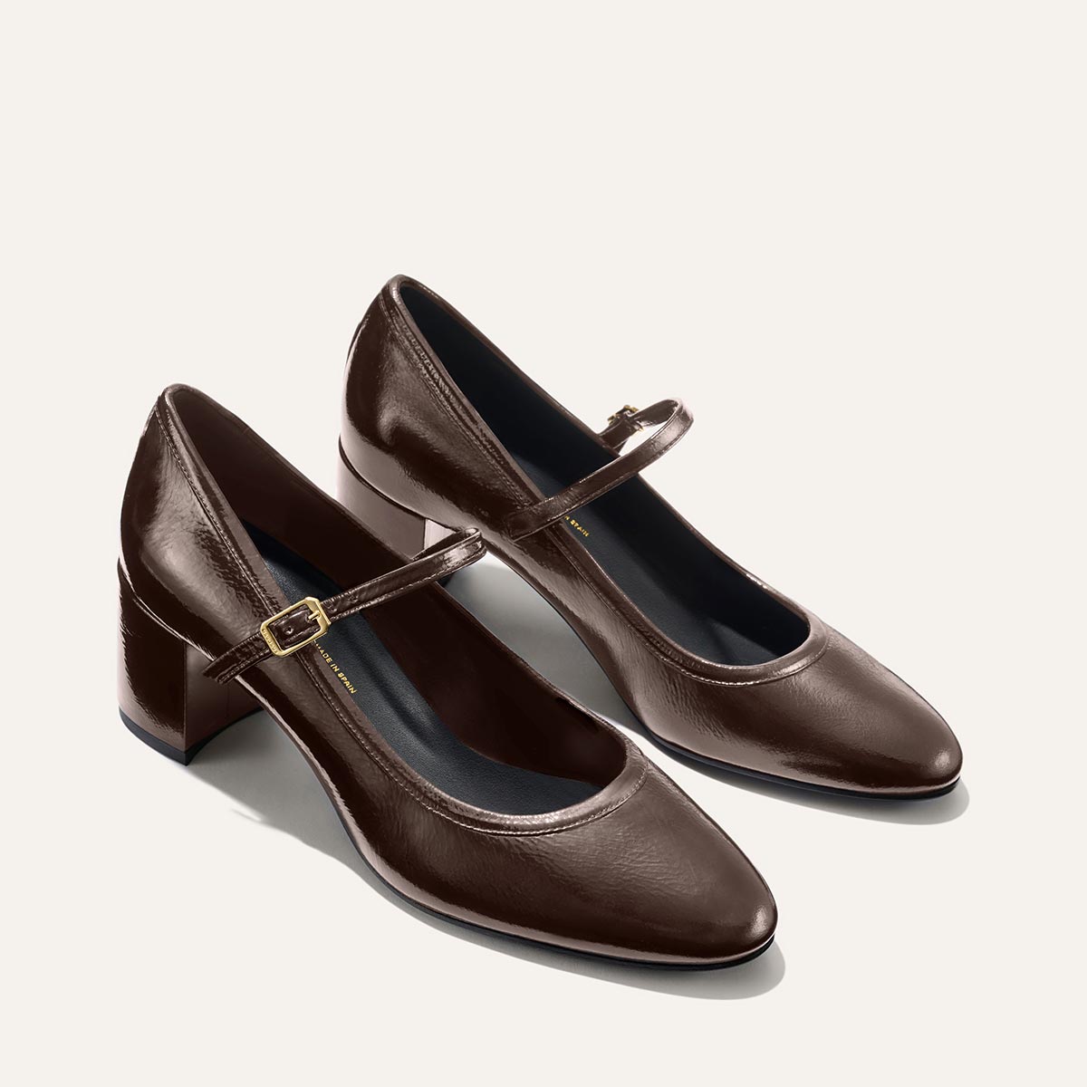 The Mira Jane - Cocoa Crinkle Patent heels by margaux-development feature a classic Mary Jane design with a strap and buckle, rounded toe, and block heel, shown on a light background.
