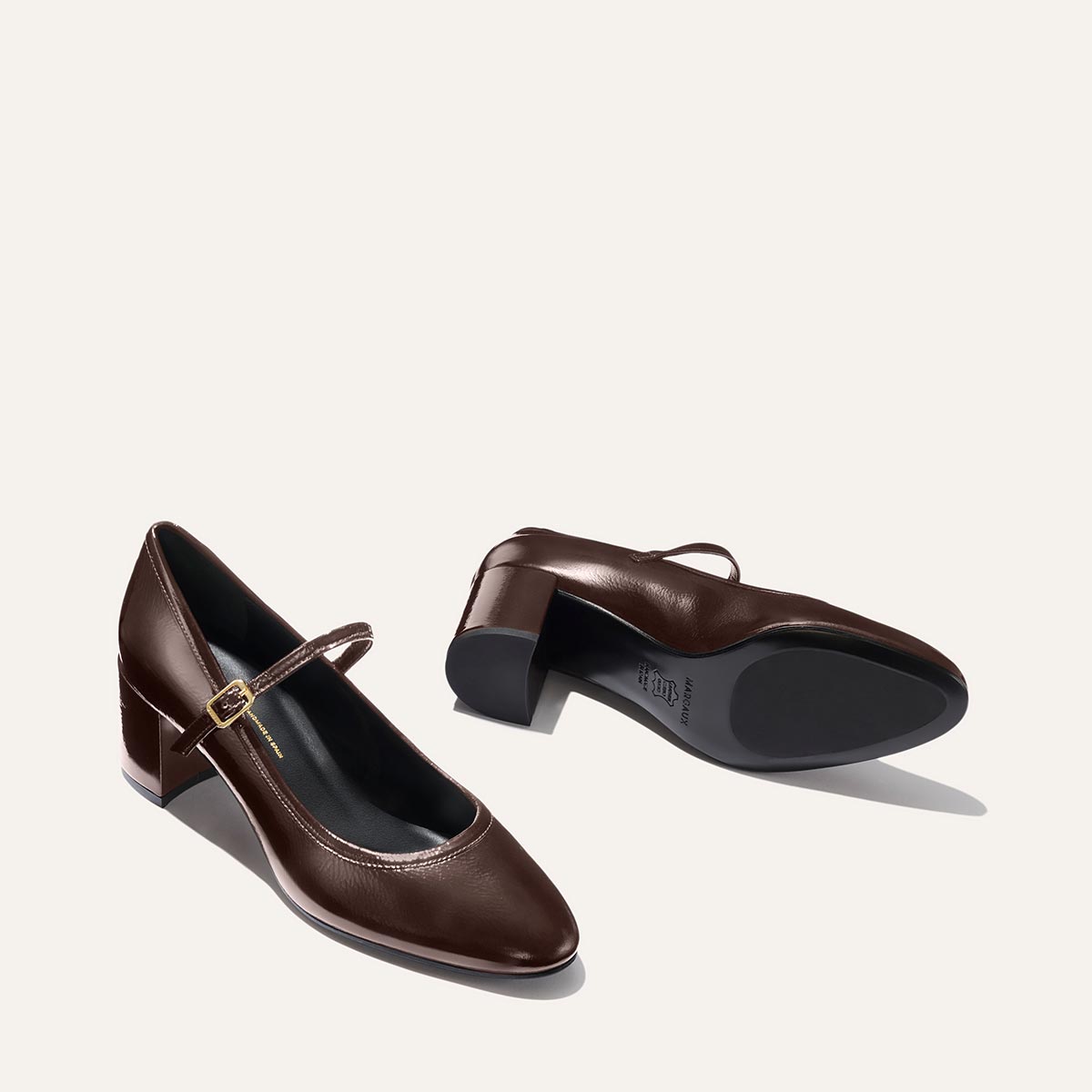 The Mira Jane - Cocoa Crinkle Patent by margaux-development features classic brown Mary Jane styling with a low block heel and a thin strap with a gold buckle, shown from the side and bottom on a light background.