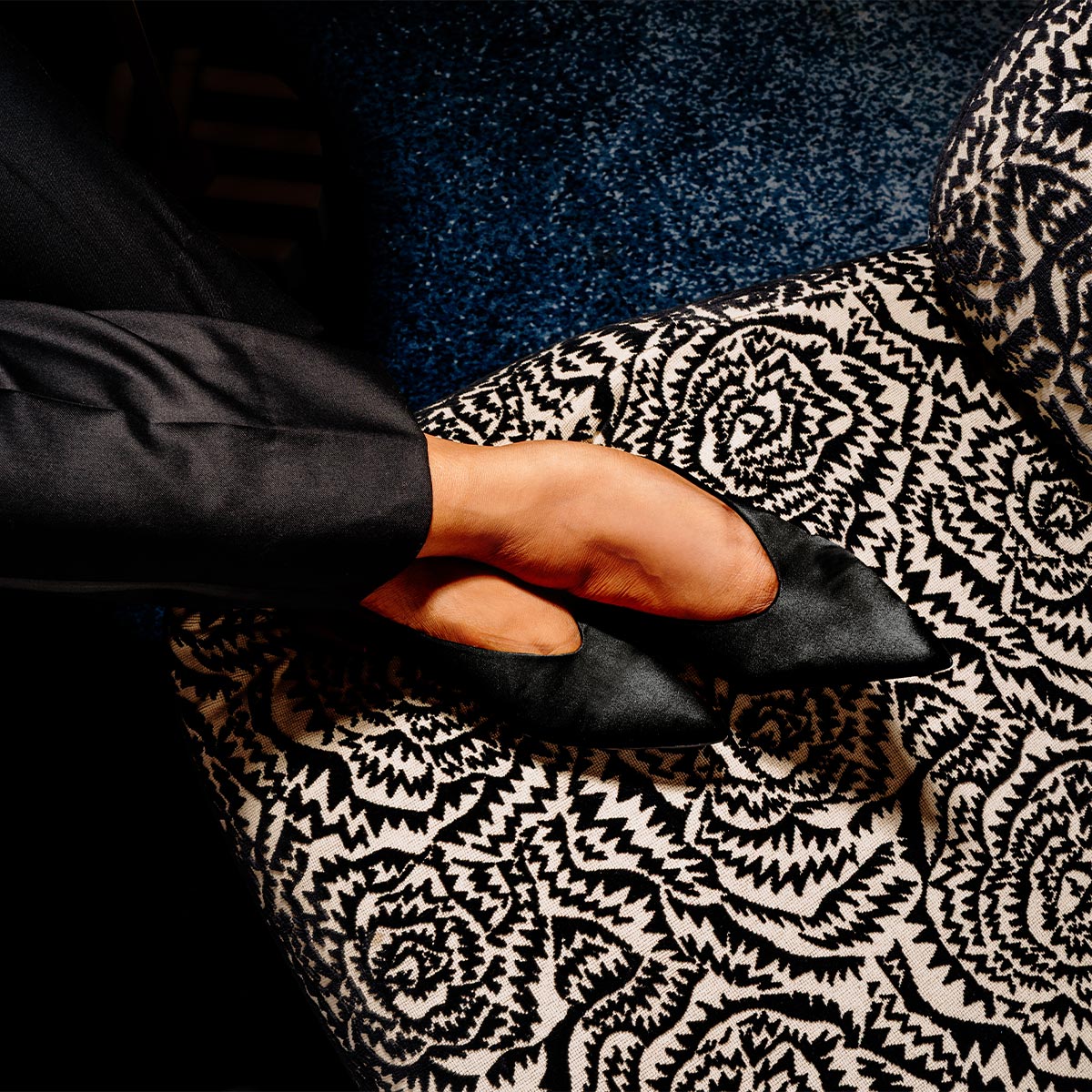 A person in dark clothing wears The Parra - Black Satin shoes by margaux-development, resting their feet on an upholstered chair with a striking black and white geometric floral pattern over a deep blue floor.