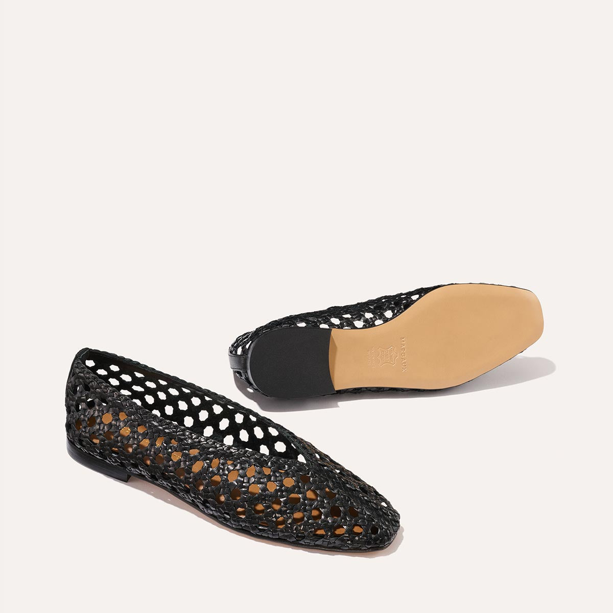 Two The Woven Paz - Black Leather flats by Margaux are displayed: one on its side to highlight the handwoven upper, the other flipped to show the tan sole, both set against a simple off-white background.