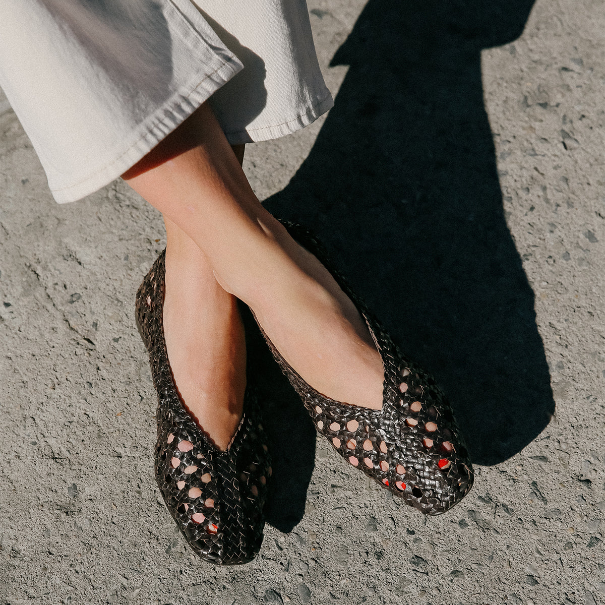 A person wearing cream-colored, high-rise pants and Margaux’s The Woven Paz flats in Espresso Leather stands on sunlit, textured concrete with red-painted toes and crossed ankles, casting a shadow.