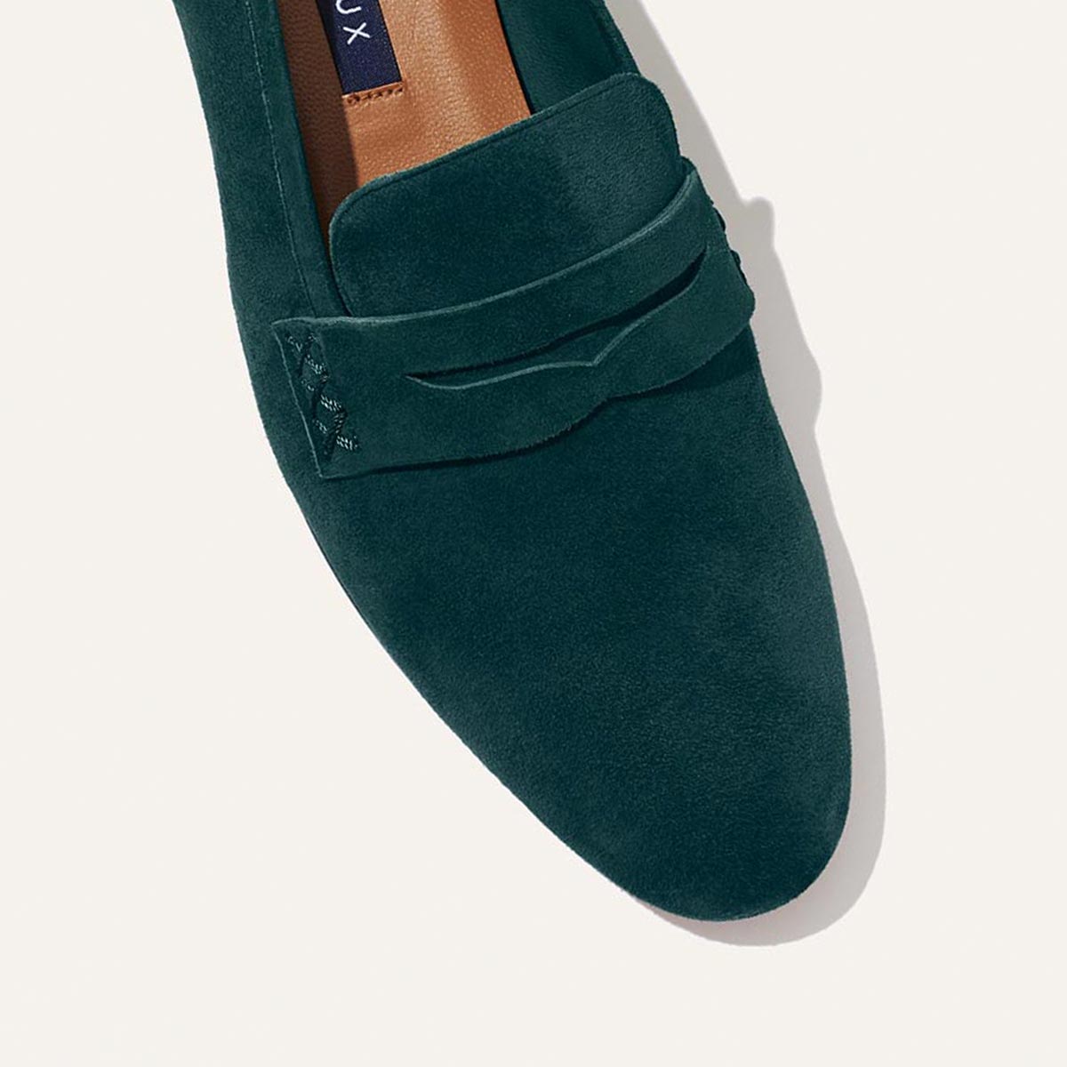 A close-up of The Penny - Emerald Suede by Margaux, a dark green Italian suede loafer with a rounded toe, penny strap detail, tan inner lining, and unlined construction for a glove-like fit, shown against a light background.
