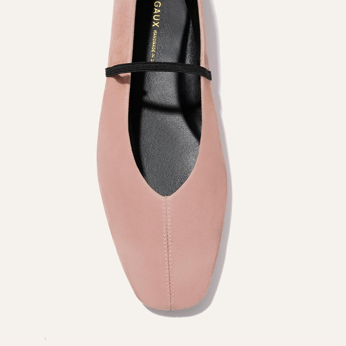 A close-up top view of The Phoebe - Dusty Rose Suede by margaux-development: a blush pink suede handmade flat with a square toe, thin black instep strap, and elegant black insole adorned with gold lettering.