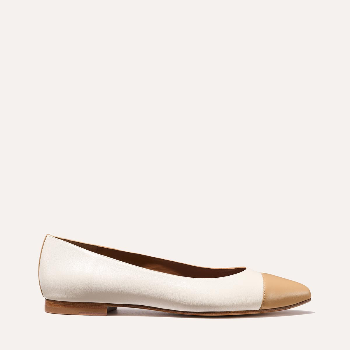 The Pointe by Margaux in Ivory and Toffee Nappa is a white flat with tan toe cap and heel, featuring a sleek silhouette and low wooden sole—shown on a plain light background.