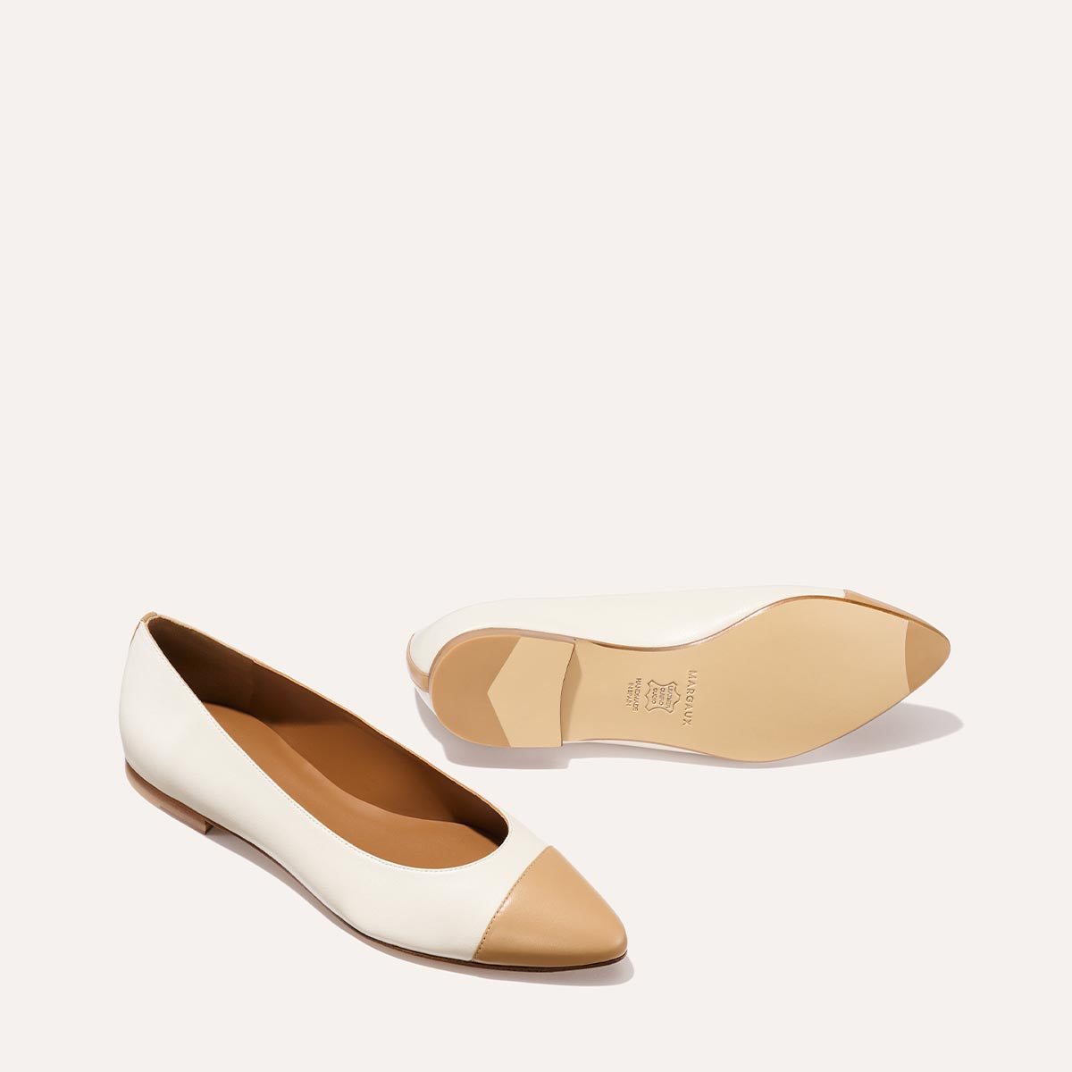 The Pointe by Margaux features a sleek, minimalist silhouette in ivory and toffee nappa. These women’s flats have cream bodies, tan pointed toe caps, low heels, and are shown upright and on their side to reveal the sole.
