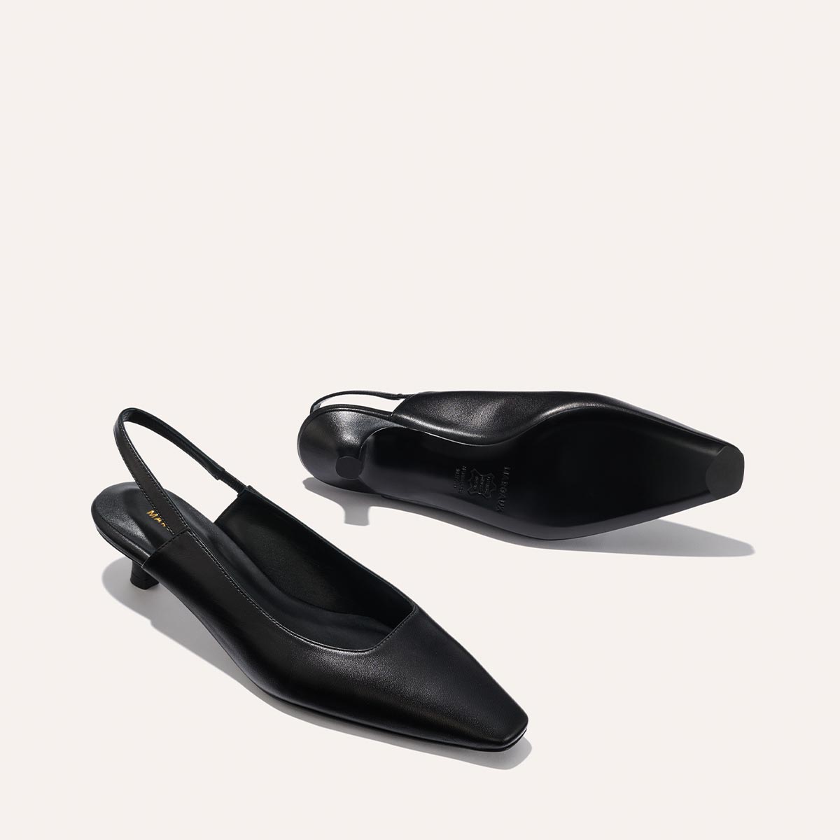 The Sofia Sling - Black Nappa by margaux-development is shown on a light background. One shoe stands upright while the other lies on its side, highlighting the sole, snip nose toe, and low micro heel design.