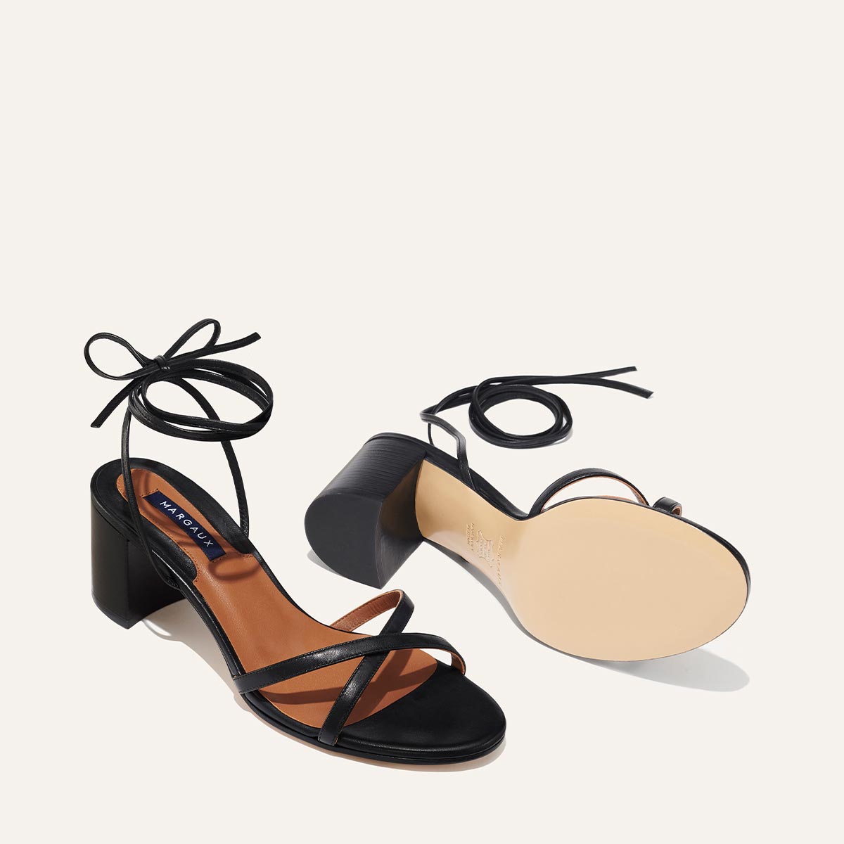 The Soho Sandal - Black Nappa by margaux-development features black wrap design, block heels, and ankle tie straps. One stands upright and the other lies on its side to show the light-colored sole—perfect for summer.