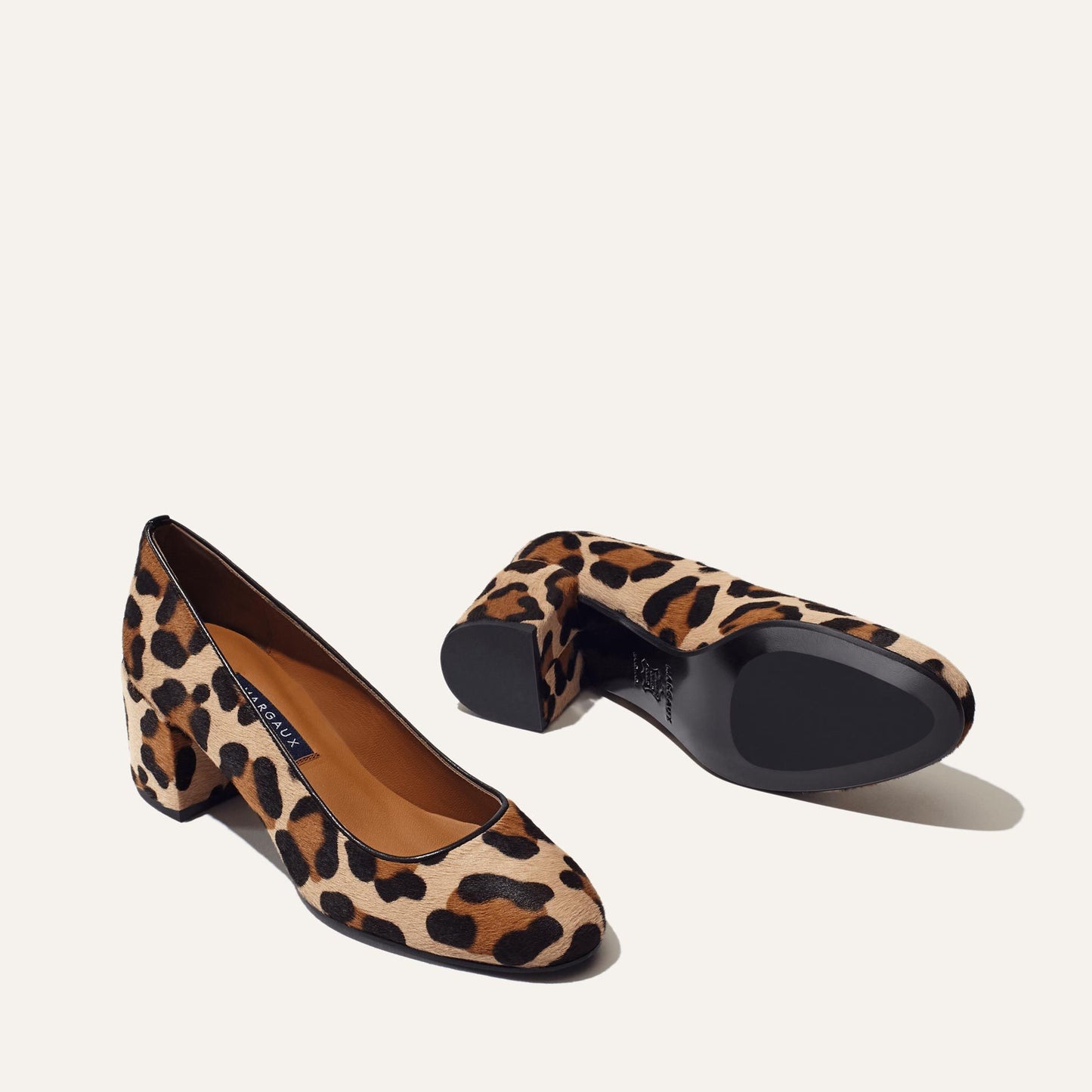 A pair of Margaux’s The Heel in Tan Leopard Haircalf is shown—one shoe upright, the other on its side showcasing the sole—set against a plain off-white background.